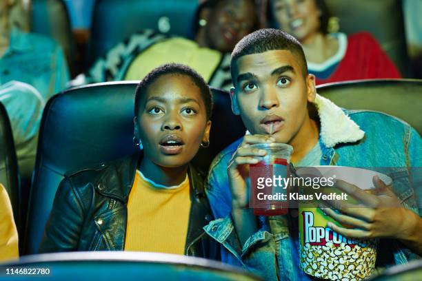 surprised young man drinking soda while watching movie with friend in cinema hall - filmindustrie stock-fotos und bilder