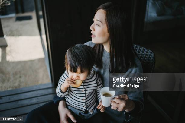cute little daughter sitting on mother's lap eating cookie while mother drinking coffee, they are relaxing on the balcony and enjoying intimate family time together - holiday resort family sunshine stock pictures, royalty-free photos & images