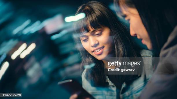multiethnic female friends share smartphone on city street at night. - differential focus stock pictures, royalty-free photos & images