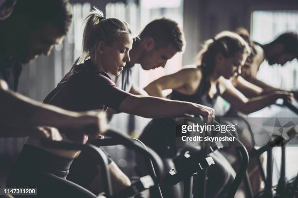 grand groupe de personnes athlétiques ayant la formation sportive sur des vélos d’exercice dans une salle de gym. - cours de spinning photos et images de collection