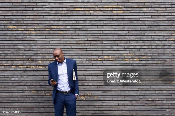 portrait of smiling businessman with newspaper wearing glasses and blue suit looking at mobile phone - england media access stock-fotos und bilder