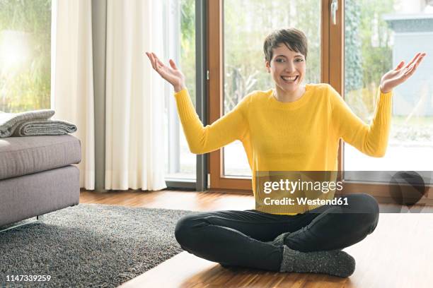 portrait of happy woman sitting on the floor of living room with hands raised - schneidersitz stock-fotos und bilder