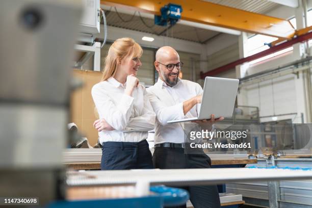 smiling businessman and businesswoman using laptop in factory - smart factory stock-fotos und bilder