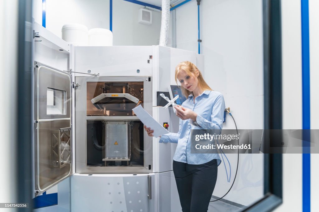 Woman holding paper and workpiece at 3d printer