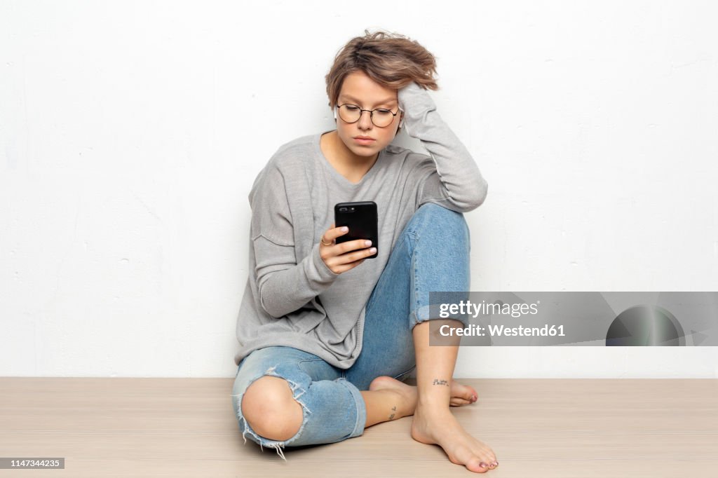 Portrait of young woman with wireless earphones sitting on the floor looking at cell phone
