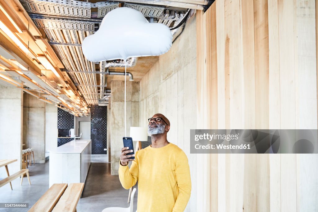 Mature businessman holding cell phone attached to cloud balloon