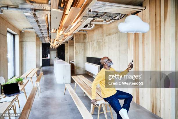 mature businessman holding cell phone attached to cloud balloon - büro und wolke stock-fotos und bilder