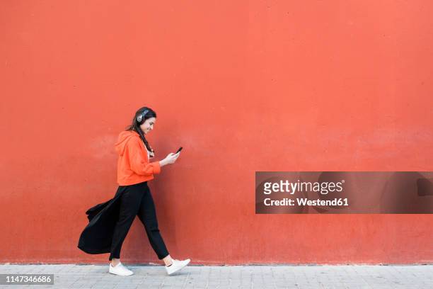 young dancer walking and using the phone and headphones in front of a red wall - hoodie headphones - fotografias e filmes do acervo