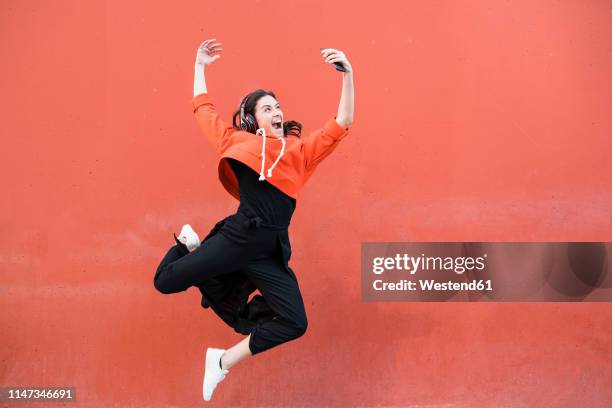 young contemporary dancer jumping and using smartphone in front of a red wall - phone mid air stock pictures, royalty-free photos & images