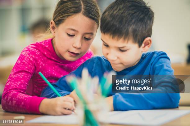 mädchen hält die hand eines klassenkameraden und hilft ihm, in seinem notizbuch zu schreiben - school boy girl stock-fotos und bilder
