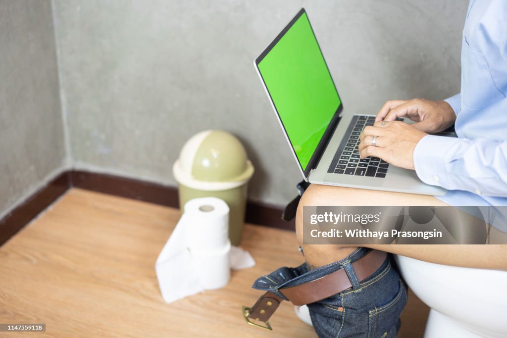 The young man is tired of using a laptop while sitting in the bathroom. The concept of hard workers