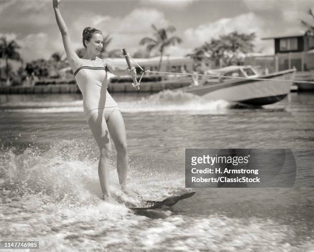 1950s EXCITED WOMAN WATER SKIING POWER BOAT SPEEDING IN BACKGROUND ON PALM TREE LINED CANAL MIAMI BEACH FLORIDA USA