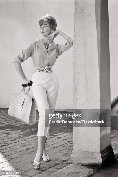 1950s 1960s FULL LENGTH PORTRAIT WOMAN WEARING BUTTON DOWN SHIRT CAPRI PANTS HOLDING STRAW BAG SUNGLASSES POSING BY COLUMN