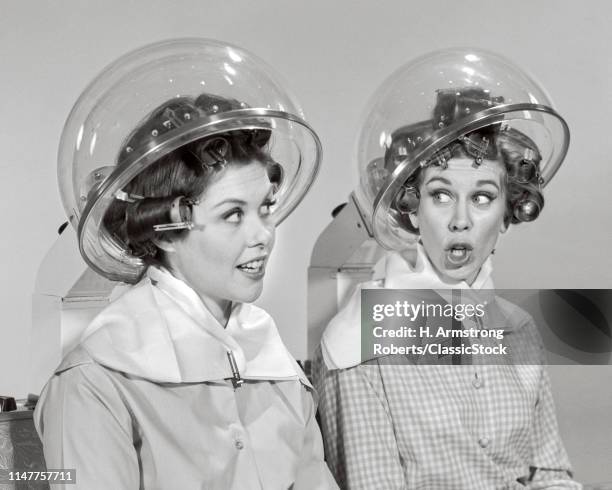 1960s Two women rollers curler in their hair sitting under hair salon dryers talking gossiping