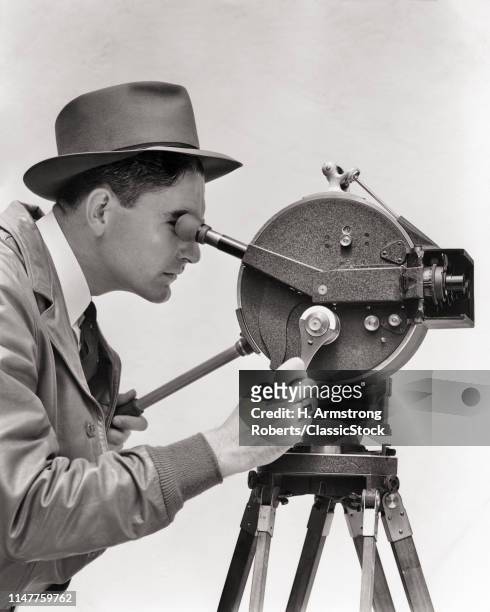 1930s PORTRAIT PROFILE MAN CINEMATOGRAPHER OPERATING AKELEY HAND CRANK PORTABLE 35 MM FILM MOVIE CAMERA MOUNTED ON TRIPOD