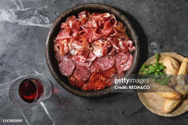 Antipasto meat platter assorti of sliced jamon, salami, chorizo sausage in terracotta tray with bread and glass of red wine over black marble...