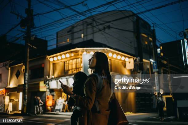 young asian mother with little daughter using mobile app on smartphone to search for direction while travelling in illuminated city street in japan at night - fukuoka city photos et images de collection