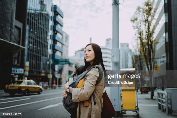 young asian mother with baby girl waiting for taxi ride in downtown city street, with urban city scene as background - madre ama de casa fotografías e imágenes de stock