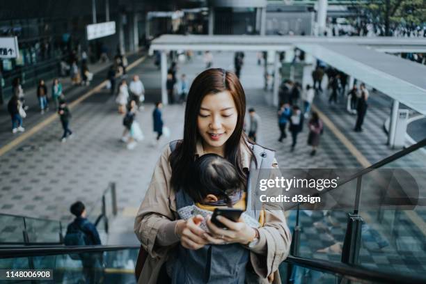 young asian mother with little daughter using smartphone while riding on escalator in downtown city, with busy commuters in the background - family mall stockfoto's en -beelden