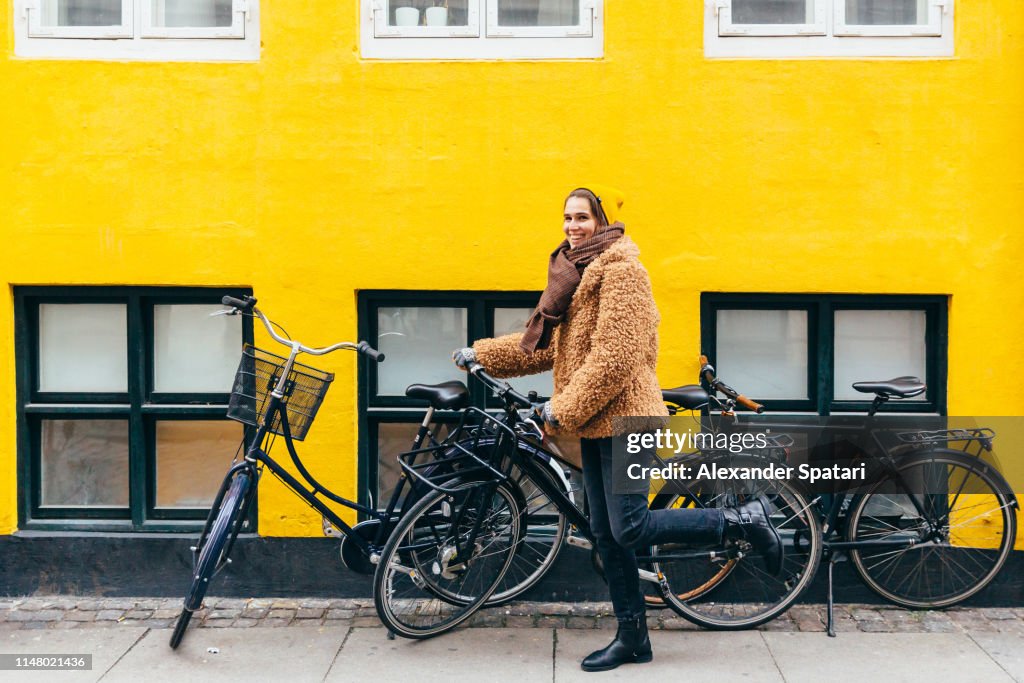 Happy young smiling woman with bicycle against yellow wall in Copenhagen