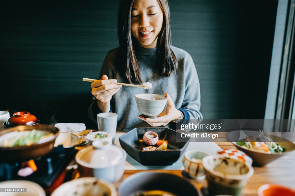 Smiling young Asian woman enjoying delicate Japanese style cuisine with various side dishes, seafood and green tea in restaurant