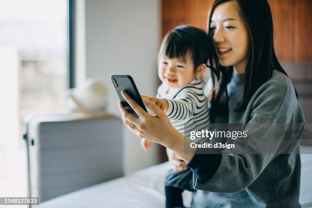 young asian mother and cute little daughter smiling joyfully while taking selfie on smartphone in hotel room during vacation - baby smartphone stock pictures, royalty-free photos & images