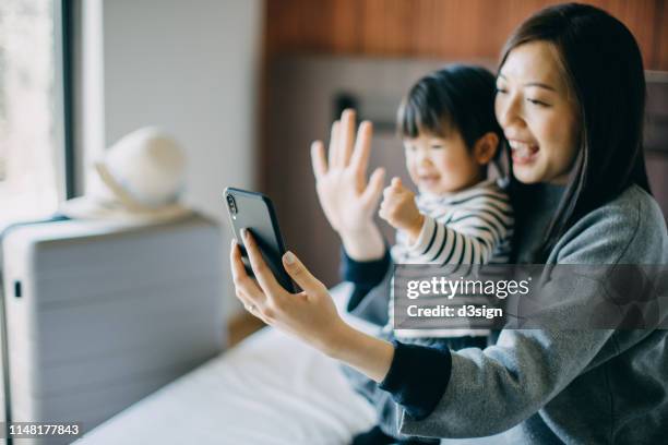 Young Asian mother and cute little daughter having video call on smartphone with family in hotel room while on vacation and smiling joyfully