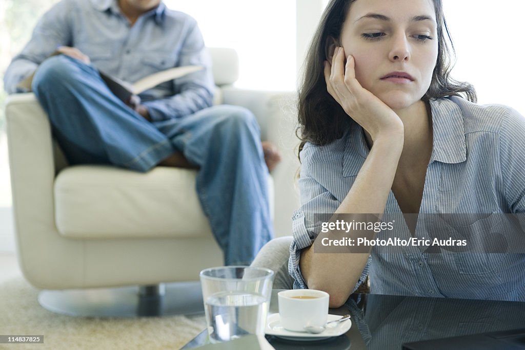 Woman leaning on elbow looking sad, man reading on sofa in background