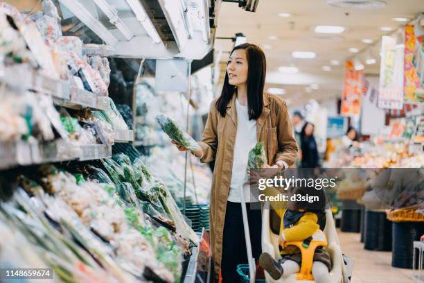 cute little daughter sitting in a shopping cart grocery shopping for fresh organic vegetables with young asian mother in a supermarket - asian family shopping stock-fotos und bilder