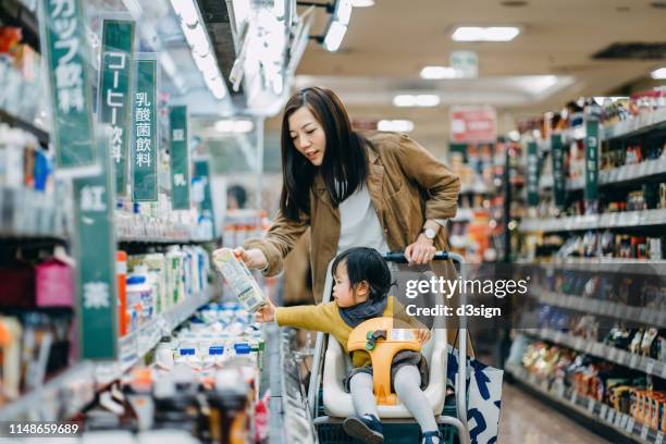 cute little daughter sitting in a shopping cart grocery shopping for dairy product with young asian mother in a supermarket - mom buying milk photos et images de collection