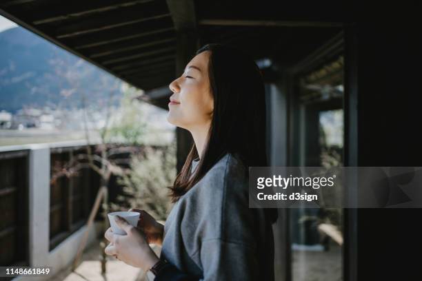 beautiful young asian woman drinking coffee and enjoying fresh air on balcony in the morning - espacio confortable fotografías e imágenes de stock