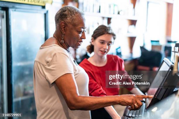 die aktive seniorin, 77-jährige, afroamerikanische geschäftsfrau, geschäftsinhaberin, die dem neuen mitarbeiter, dem 18-jährigen kaukasischen weißen mädchen, beigebracht hat, wie man die computerisierte kasse in dem kleinen lokal nutzt. - black woman on cash register stock-fotos und bilder