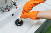 Person in protective orange gloves unblocking a clogged sink with plunger or rubber pump. Close-up, selective focus.