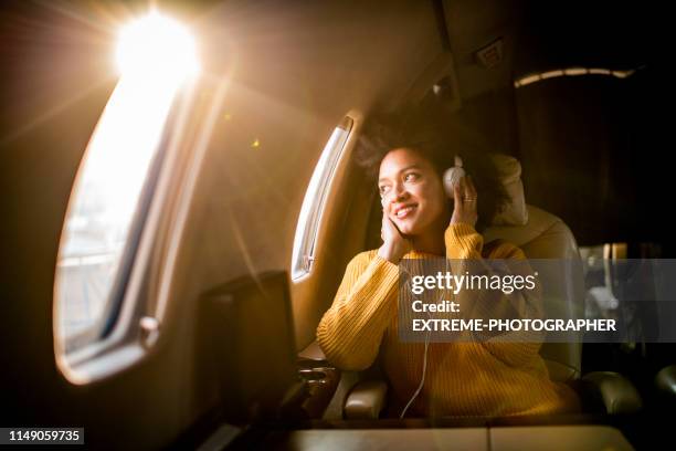 joven mujer moderna sentada en un jet privado, escuchando música a través de los auriculares y mirando a través de la ventana - air travel fotografías e imágenes de stock