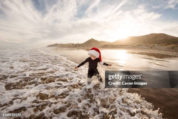toddler boy wearing red hat splashing in waves at beach at dusk - christmas travel stock pictures, royalty-free photos & images