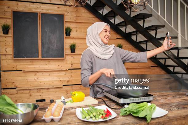 eine junge malaiische muslimis, die sich beim streaming befindet, während sie in der küche kochen - malay culture stock-fotos und bilder