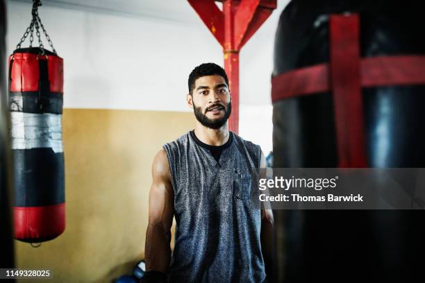 portrait of male boxer standing by heavy bag in boxing gym - people portraits hobbies stockfoto's en -beelden