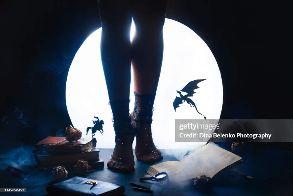 A young female writer with a fairy tale book. Dragon and Knight silhouettes against shiny Moon background. Legs in starry socks