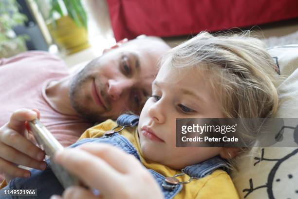 father and young daughter laying down, looking at phone - children screen stock pictures, royalty-free photos & images