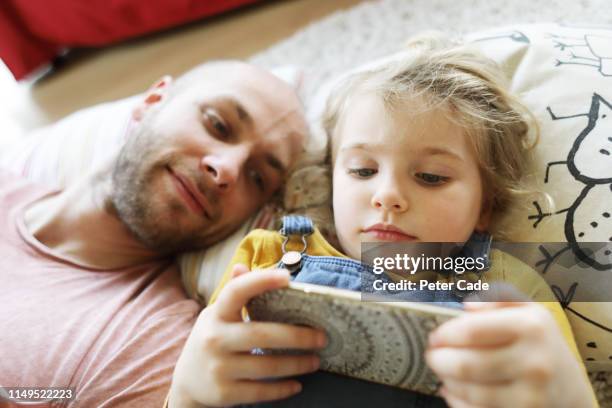 father and young daughter laying down, looking at phone - family time stock pictures, royalty-free photos & images