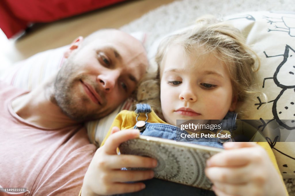 Father and young daughter laying down, looking at phone