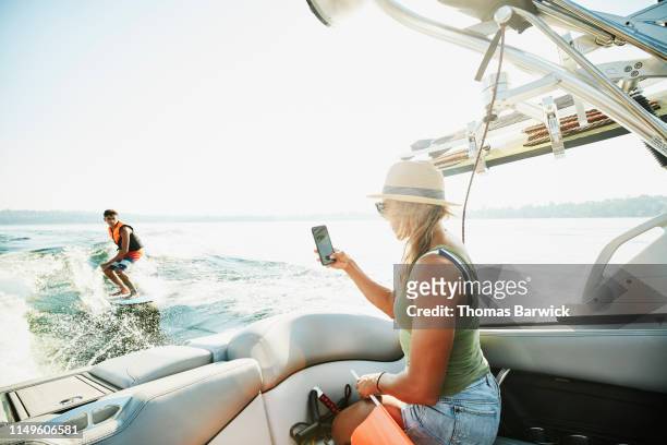 mother taking photo with smart phone of teenage son wake surfing behind boat on summer afternoon - woman surfing stock pictures, royalty-free photos & images