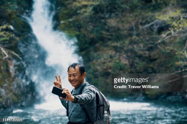 Senior man taking a selfie with a smart phone in front of a waterfall while hiking in a forest