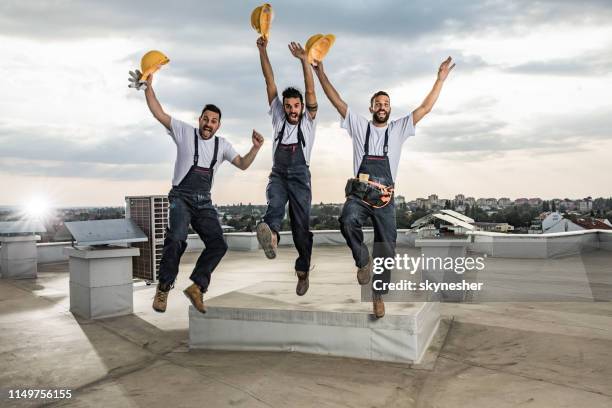 fröhliche bauarbeiter haben spaß beim springen mit den auf einem dach aufgezogenen armen. - handwerker stock-fotos und bilder