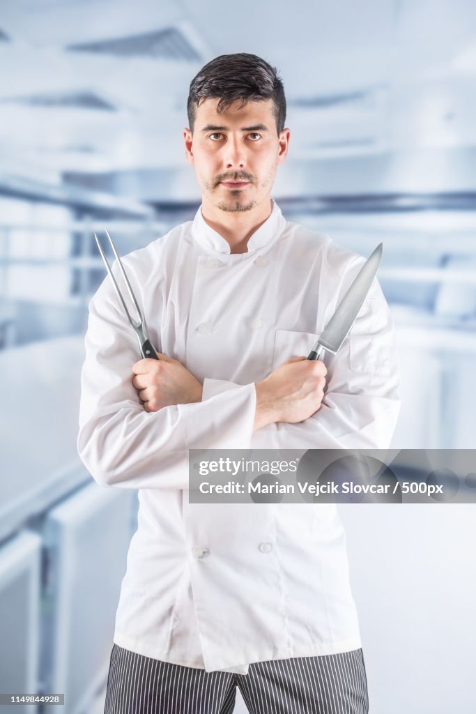 Chef Professional In Restaurant Kitchen Standing With Crossed Hands And Holding Fork And Knife