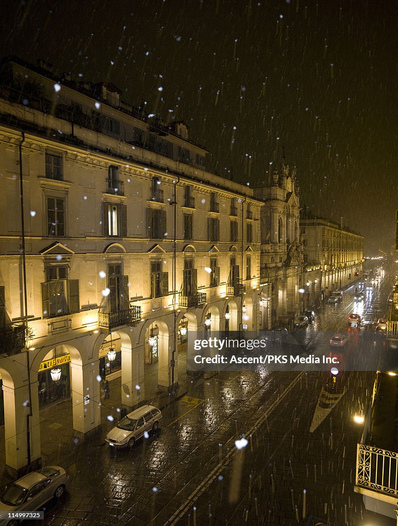 Snow falls on city street at night, Turin