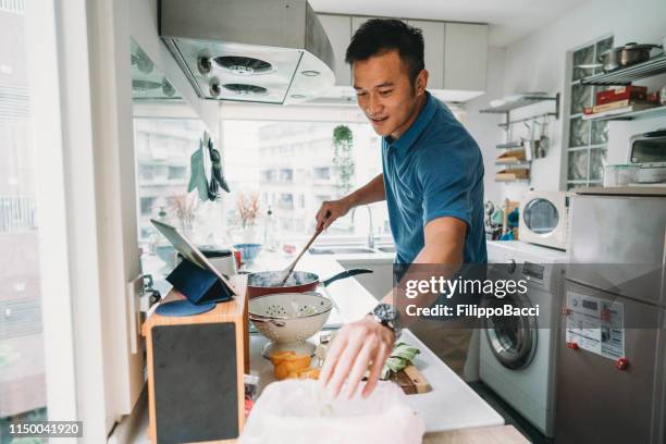 man förbereder middag i köket tittar på ett recept på tabletten - asian man cooking bildbanksfoton och bilder