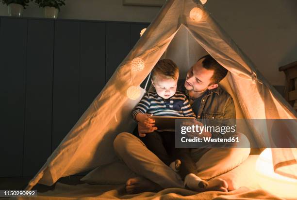 father and son sharing a tablet in a dark tent at home - family livingroom stock-fotos und bilder