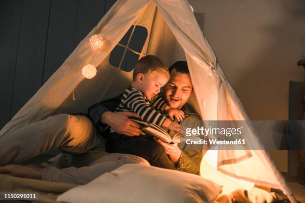 father reading book to son at an illuminated tent at home - zelt nacht stock-fotos und bilder