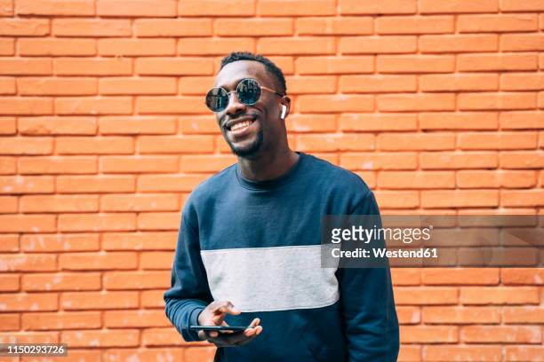 portrait of happy man wearing sunglasses listening music with wireless earphones and smartphone - young man listening to music on smart phone outdoors stockfoto's en -beelden